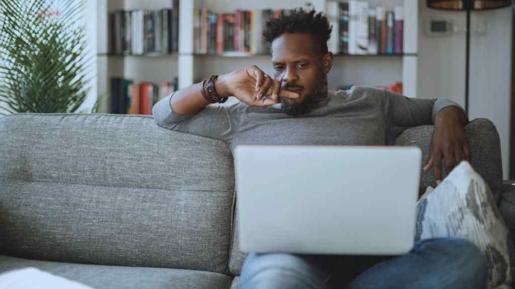 A man sits thoughtfully on a gray sofa, focused on a laptop. Behind him are shelves filled with books. The room feels calm and contemplative.