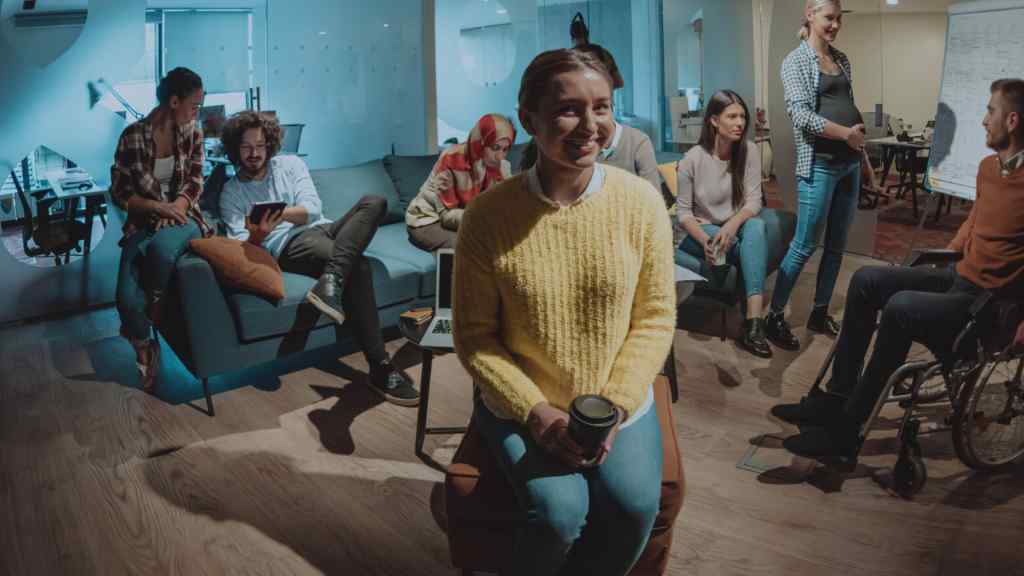 A diverse group of people in a modern office lounge, smiling and chatting. One person in the foreground wears a yellow sweater, holding a coffee cup. The mood is relaxed and collaborative.