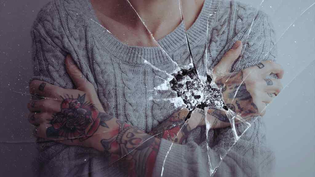 A person in a gray cable-knit sweater, arms crossed, displays colorful tattoos. The image features a cracked glass effect, conveying a sense of vulnerability.