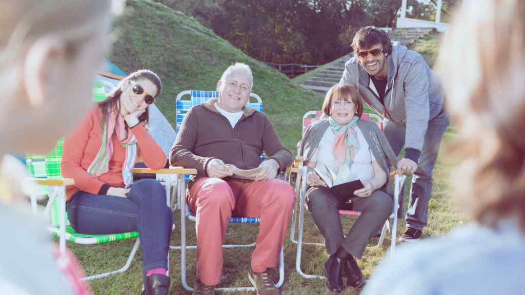 A group of four people sit outdoors, smiling and relaxed on colorful lawn chairs. They're gathered on a sunny day, conveying warmth and joy.