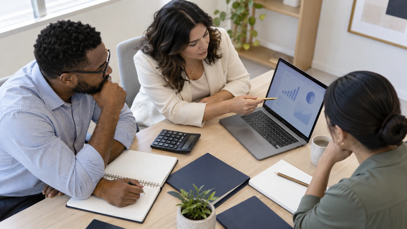 Three colleagues review a laptop chart with a calculator and notebooks on a light wood table.