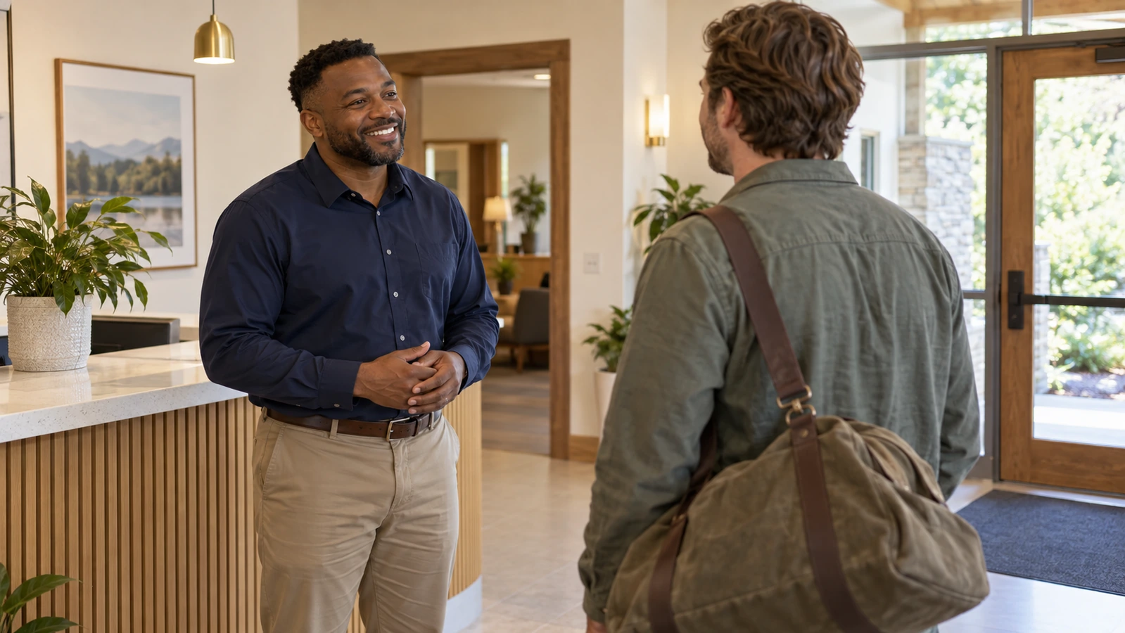 A coordinator warmly greets an adult carrying an overnight bag in a bright reception area.